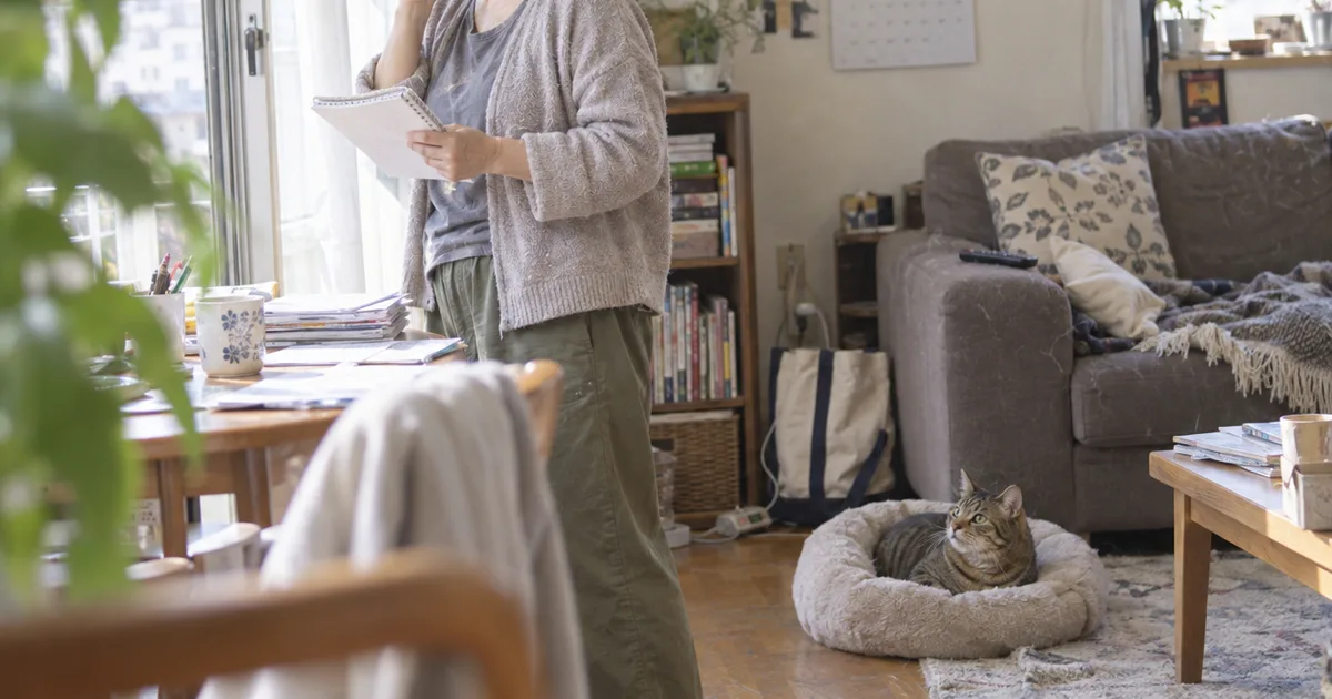 リビングでメモ帳を手にする40代の女性と、ペットベッドから見上げるキジトラ猫